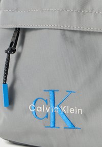 Γκρι υφασμάτινο σακίδιο πλάτης με κλείσιμο με φερμουάρ, μαύρο κορδόνι και μπλε κεντημένο λογότυπο «Calvin Klein» με χαρακτηριστική πινελιά «CK».