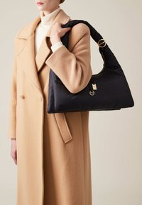Black padded handbag with a curved strap and gold hardware detail, held by a person in a beige coat against a neutral background.