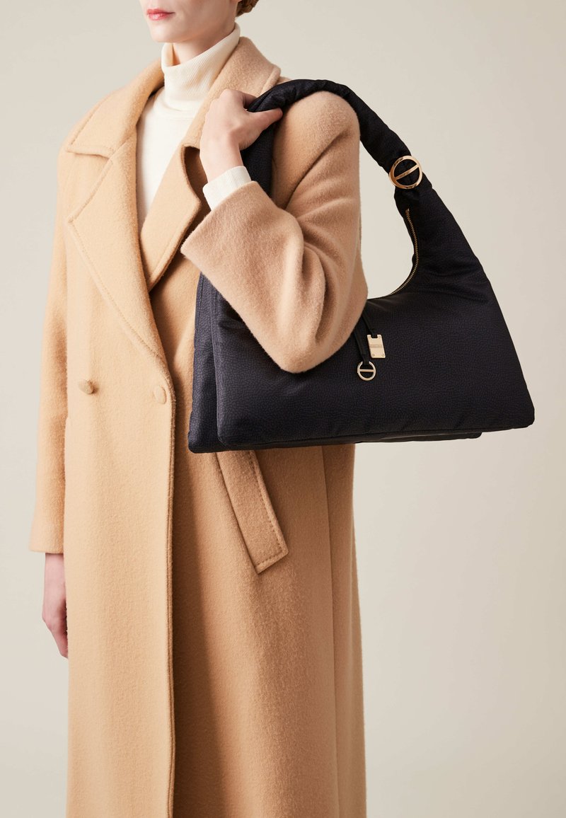 Black padded handbag with a curved strap and gold hardware detail, held by a person in a beige coat against a neutral background.