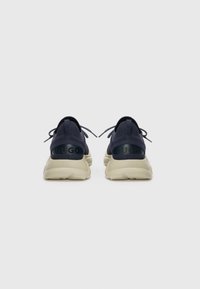 Marineblå sneakers i tekstil med en polstret kremfarget såle. Funksjoner inkluderer snøring, en logo-patch på hælen, og et strukturert overdelsdesign for bedre grep.