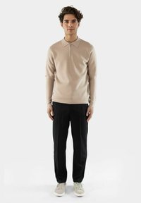 Beiger Strickpullover mit einem Reißverschlusskragen, schwarzen, taillierten Hosen und hellen Sneakers. Glatte Textur und figurbetonter Schnitt.