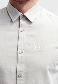 Light gray button-up shirt with a textured, striped pattern, featuring a spread collar and embroidered white logo on the left chest.