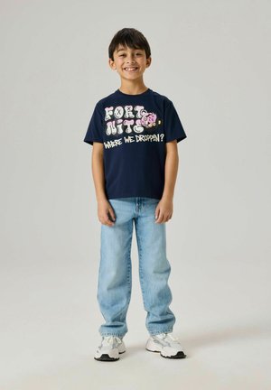 Uśmiechnięty chłopiec stojący, w granatowej koszulce z nadrukiem z kreskówką i tekstem, jasnoniebieskich dżinsach oraz białych trampkach na jednolitym tle.
