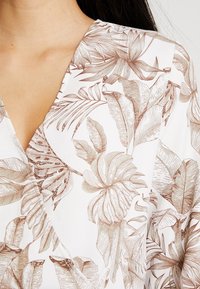 Blusa in tessuto bianco con motivo floreale, caratterizzata da illustrazioni botaniche marroni; design a scollo a V con morbide drappe e dettagli.