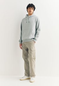 Sweat à capuche en polaire grise avec une poche avant, associé à un pantalon cargo beige avec poches latérales et des baskets blanches à semelle jaune.