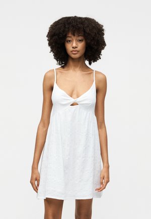 Γυναίκα με κατσαρά μαλλιά που φοράει αμάνικο λευκό φόρεμα με στριφτό μπροστινό μέρος και μικρή λεπτομέρεια με άνοιγμα, στέκεται μπροστά σε απλό φόντο.