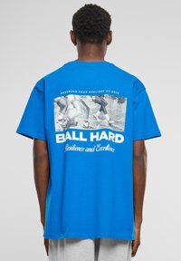 Modré bavlněné tričko s krátkými rukávy. Na zadní straně se nachází černobílý grafický potisk s textem "BALL HARD" a "Odolnost a dokonalost."