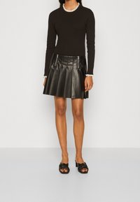 Svart långärmad topp med volangkrage, kombinerad med en svart plisserad läderkjol med dragkedjedetaljer, kompletterad med svarta sandaler.