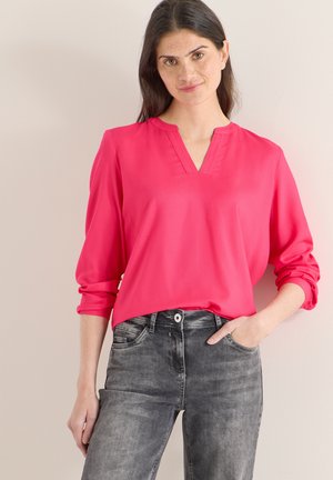 Blouse rose vif avec un décolleté en V et des manches longues retroussées, associée à un jean denim gris. Tissu lisse avec un léger éclat.