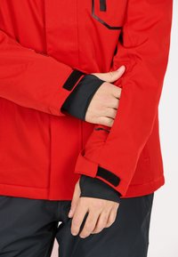 Rote Jacke mit einer glatten Textur, verstellbaren Klettverschluss-Manschetten und einer Reißverschlusstasche am Ärmel mit schwarzen Akzenten.