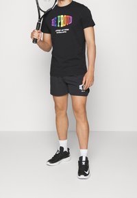 Czarna koszulka z kolorowym napisem "BĄDŹ DUMNY" oraz "PRZECIW WSZELKIM NORMOM BJÖRN BORG." Czarne szorty z białymi akcentami. Trzyma rakietę tenisową. Czarne buty.