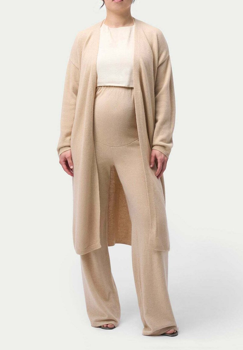 Beige stickad kavaj över en matchande topp och vida byxor. Mjuk textur, öppen front och avslappnad passform. Avslappnad design för komfort.