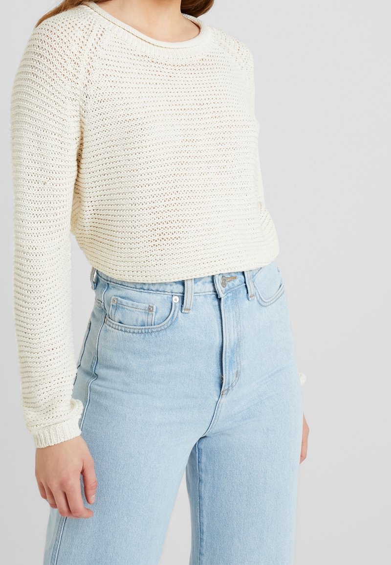 Femme portant un pull en crochet crème à manches longues associé à un jean taille haute bleu clair.