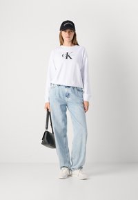 Femme portant un sweatshirt blanc Calvin Klein, un jean bleu clair, des baskets blanches et une casquette noire Calvin Klein, tenant un sac à main noir devant un fond blanc.