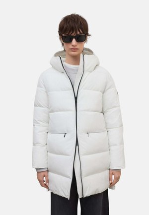 Persona que lleva gafas de sol negras y una chaqueta acolchada blanca con capucha, de pie frente a un fondo claro y sencillo.
