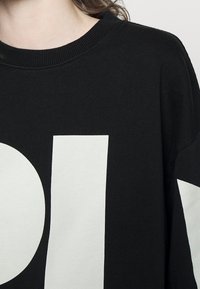 Personne portant un sweat-shirt noir avec de grandes lettres blanches en bloc partiellement visibles sur le devant et la manche.