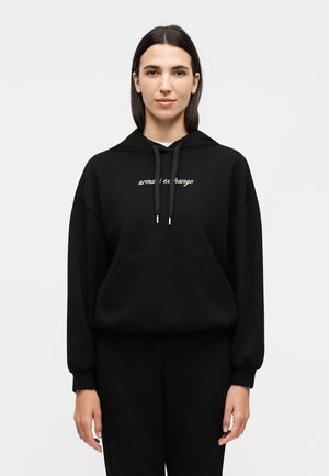 Kobieta z prostymi ciemnymi włosami nosi czarną bluzę z kapturem z przednią kieszenią oraz białym napisem „armani exchange” na piersi, stojąc na jednolitym tle.