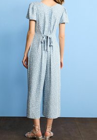 Mujer con un mono de flores azul claro de mangas cortas y cintura anudada, de pie frente a una pared azul con sandalias plateadas.