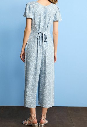Mujer con un mono de flores azul claro de mangas cortas y cintura anudada, de pie frente a una pared azul con sandalias plateadas.
