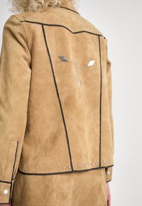 Giacca in suede marrone con borchie argento e dettagli a forma di diamante sul retro, con cuciture nere che delineano il design.