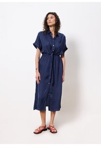 Navyblaue, taillierte Hemdkleid aus Satin mit kurzen, hochgekrempelten Ärmeln und einem Bindegürtel in der Taille. Getragen mit roten Sandalen.