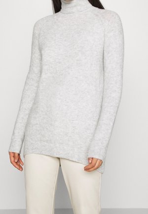 Personne portant un pull à col roulé duveteux gris clair à manches longues et un pantalon écru sur un fond blanc uni.