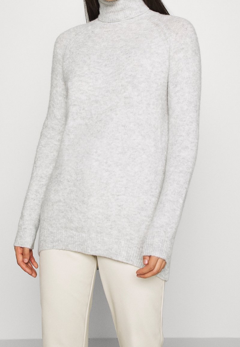 Personne portant un pull à col roulé duveteux gris clair à manches longues et un pantalon écru sur un fond blanc uni.
