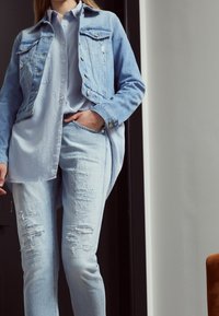 Chaqueta de mezclilla azul clara con botones plateados, combinada con una camisa azul clara y jeans desgastados de mezclilla azul clara. Textura suave, corte ajustado.
