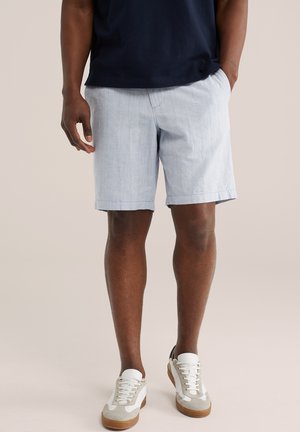 Mann trägt hellblaue Shorts, dunkelblaues Hemd und weiße Turnschuhe und steht vor einem schlichten beigen Hintergrund.