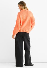 Jasnoróżowy, oversize dzianinowy sweter z prążkowanym dołem i luźnymi rękawami, w zestawieniu z szerokimi czarnymi dżinsami i czarnymi butami.