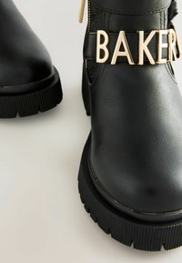 Čierne boty na členku z faux kože s robustnou gumovou podrážkou, vybavené zlatým emblémom "BAKER" na remienku a štruktúrovaným detailom na hornej časti.