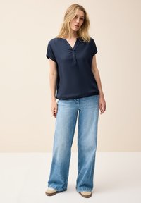 Blusa corta blu navy con bottoni, abbinata a jeans a gamba larga blu chiaro e sneaker beige. Tessuto morbido e vestibilità comoda.