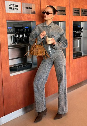 Woman in textured gray jacket and pants with brown bag and boots holds cup and spoon, standing by frozen yogurt dispensers with flavor labels.