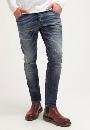 Strak zittende, versleten blauwe spijkerbroek gecombineerd met bordeauxrode leren Chelsea boots en een wit shirt met lange mouwen, handen in de zakken.