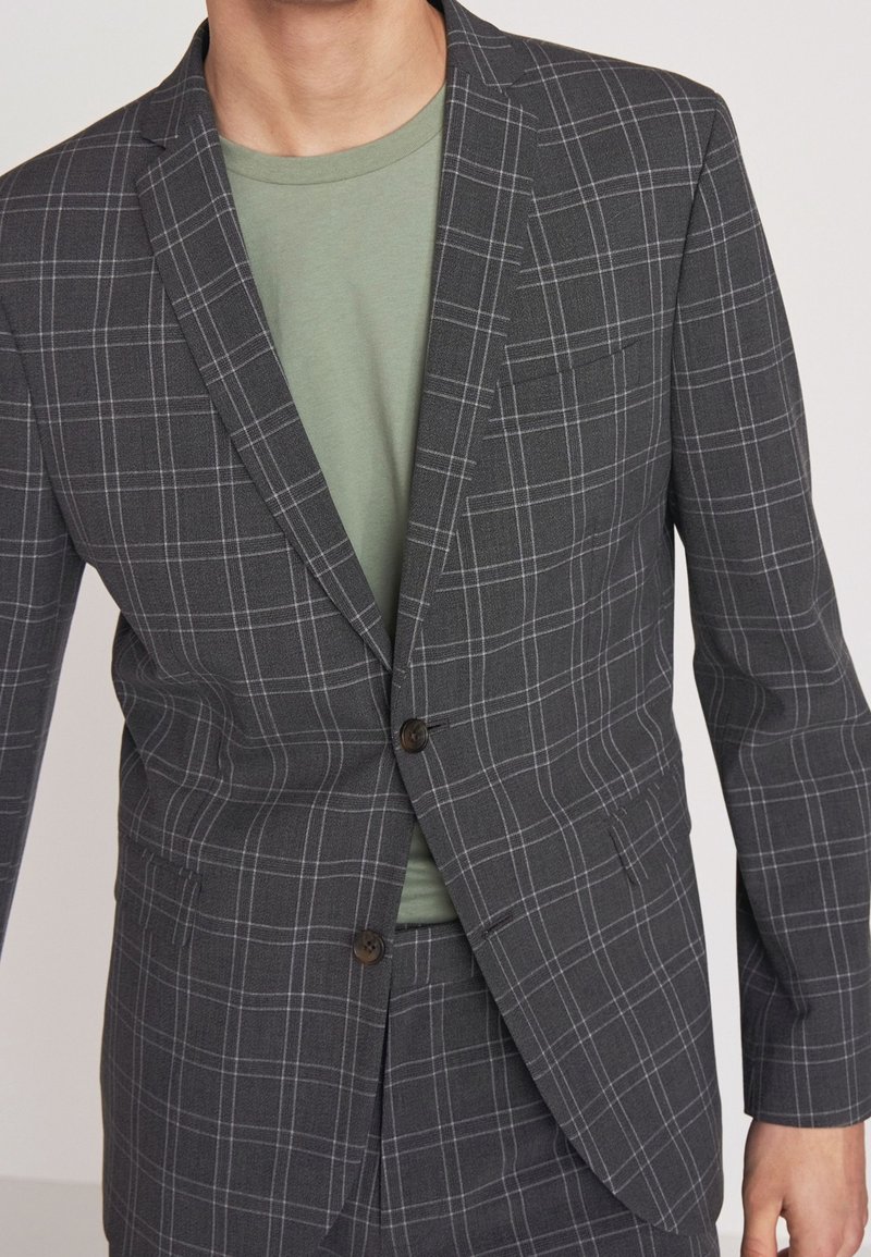 Blazer de color gris oscuro con un patrón de cuadrícula, que presenta una solapa con muesca, cierre de dos botones y un bolsillo. Llevado sobre una camisa verde.