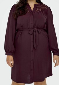 Bordeauxfärgad klänning med långa ärmar och knapprad framtill, med spetsdetalj vid halsringningen och ett knytbart midjebälte. Mjuk, texturerad tyg.