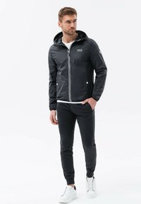 Sort vandtæt jakke med hætte, lynlåslukning, hvide accenter, båret over en hvid skjorte og sorte tapered bukser, parret med hvide sneakers.