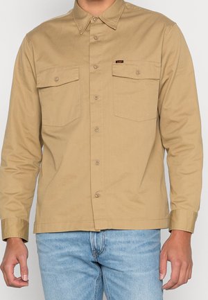 Mężczyzna w beżowej koszuli na guziki z dwoma przednimi kieszeniami na klapie i jasnoniebieskich dżinsach, stojący na jednolitym tle.