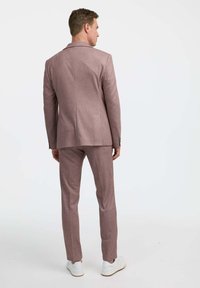 Világos rózsaszín, testre szabott szabású öltöny, slim-fit dizájnnal, texturált anyagból, kilapított hajtókával, fehér sportcipővel, hátulról nézve.