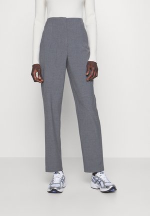 Person iført højttaljede grå bukser med lige ben, hvid ribbet langærmet top og hvide atletiske sneakers står på en neutral baggrund.