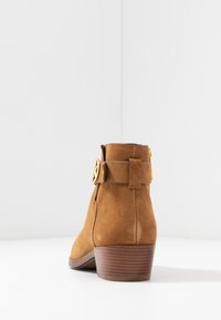 Brown suede ankle boot with a stacked heel, featuring a decorative gold buckle strap and smooth rounded toe.