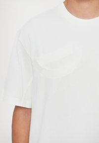 Weißes Baumwoll-T-Shirt mit Rundhalsausschnitt und kurzen Ärmeln, ausgestattet mit einer Brusttasche und dezent geprägtem Logo auf der Vorderseite.