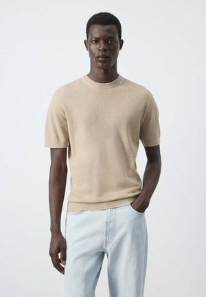 Jeune homme portant un pull beige à manches courtes et un jean bleu clair, debout avec une main dans la poche sur un fond uni.