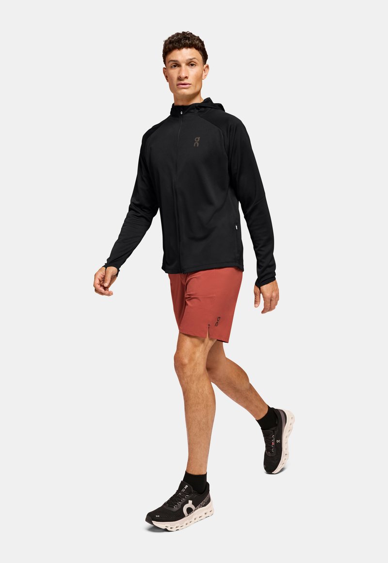Schwarze Zip-Jacke mit Kapuze, kombiniert mit roten Shorts und schwarzen Schuhen mit sichtbarer weißer Sohle. Das Model geht.