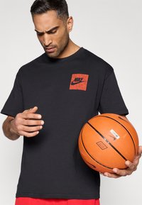 Μαύρο βαμβακερό μπλουζάκι με κόκκινο τετράγωνο λογότυπο Nike στο στήθος. Το μοντέλο κρατάει μια πορτοκαλί μπάλα του μπάσκετ με μαύρες λεπτομέρειες στο δεξί του χέρι.