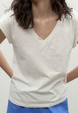 Koszulka z dekoltem w serek z miękkiej bawełny w kremowym kolorze, z krótkimi rękawami i luźnym krojem. Mały czerwony detal w postaci logo blisko dolnego brzegu.