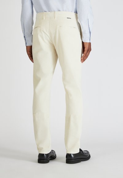 Jasnokolorowe spodnie z cordurowej tkaniny o slim fit, z tylnymi kieszeniami, w połączeniu z czarnymi skórzanymi butami. Materiał ma pionową teksturę prążków.