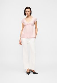 Γυναίκα που φοράει ένα ανοιχτό ροζ μπλουζάκι με κοντά μανίκια και λεπτομέρεια ρίγας, καθώς και λευκό παντελόνι φαρδιάς γραμμής, στέκεται με το ένα χέρι στην τσέπη και φοράει μαύρα τακούνια.