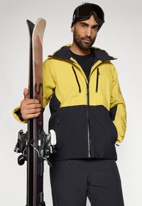 Geltonai juoda slidinėjimo striukė su užtrauktuku, reguliuojama kupra ir keliais kišenėmis. Modelis laiko slidinėjimo slides su juodais ryšiais.