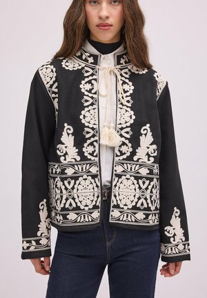 Femme portant une veste noire avec une broderie florale blanche complexe et des liens à pompons sur une chemise blanche et un jean bleu foncé.